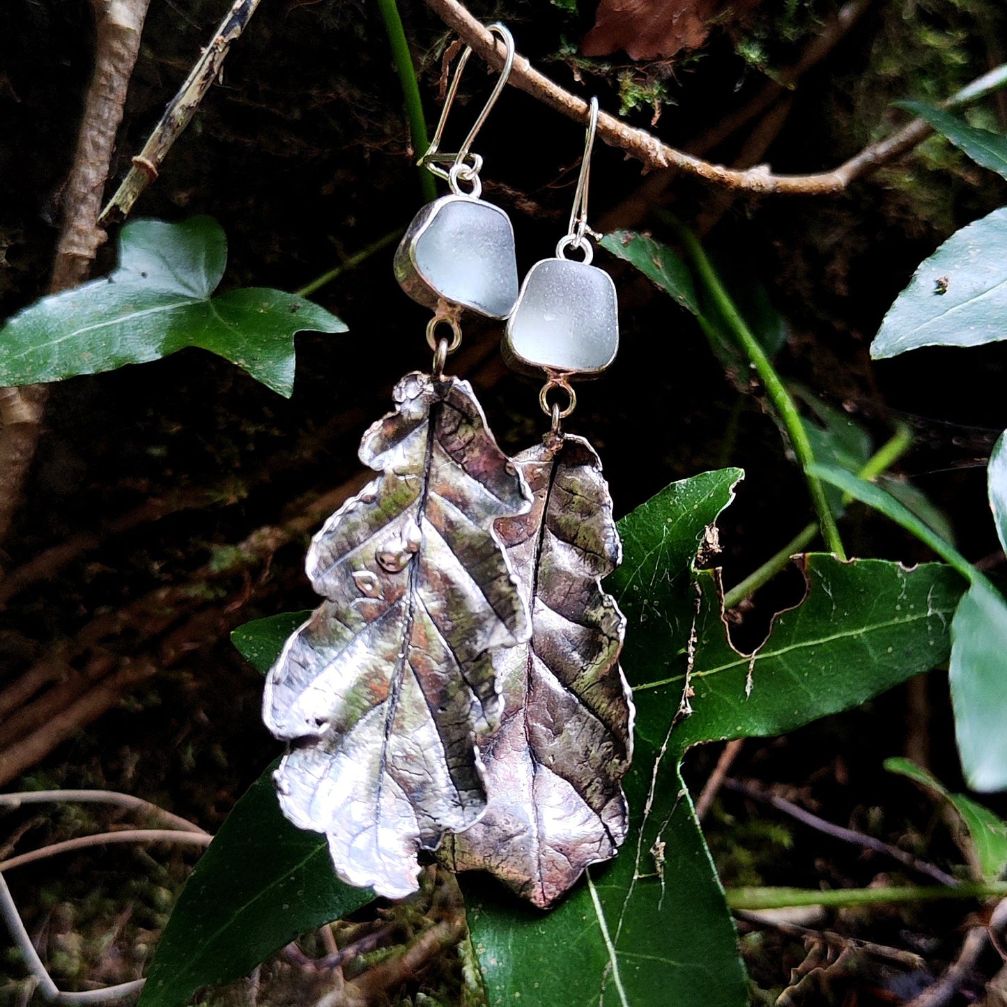 Pair of earrings with icy blue sea glass and  leaf design hanging on a branch with green leaves