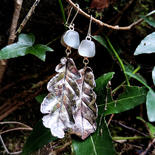 Pair of earrings with icy blue sea glass and  leaf design hanging on a branch with green leaves