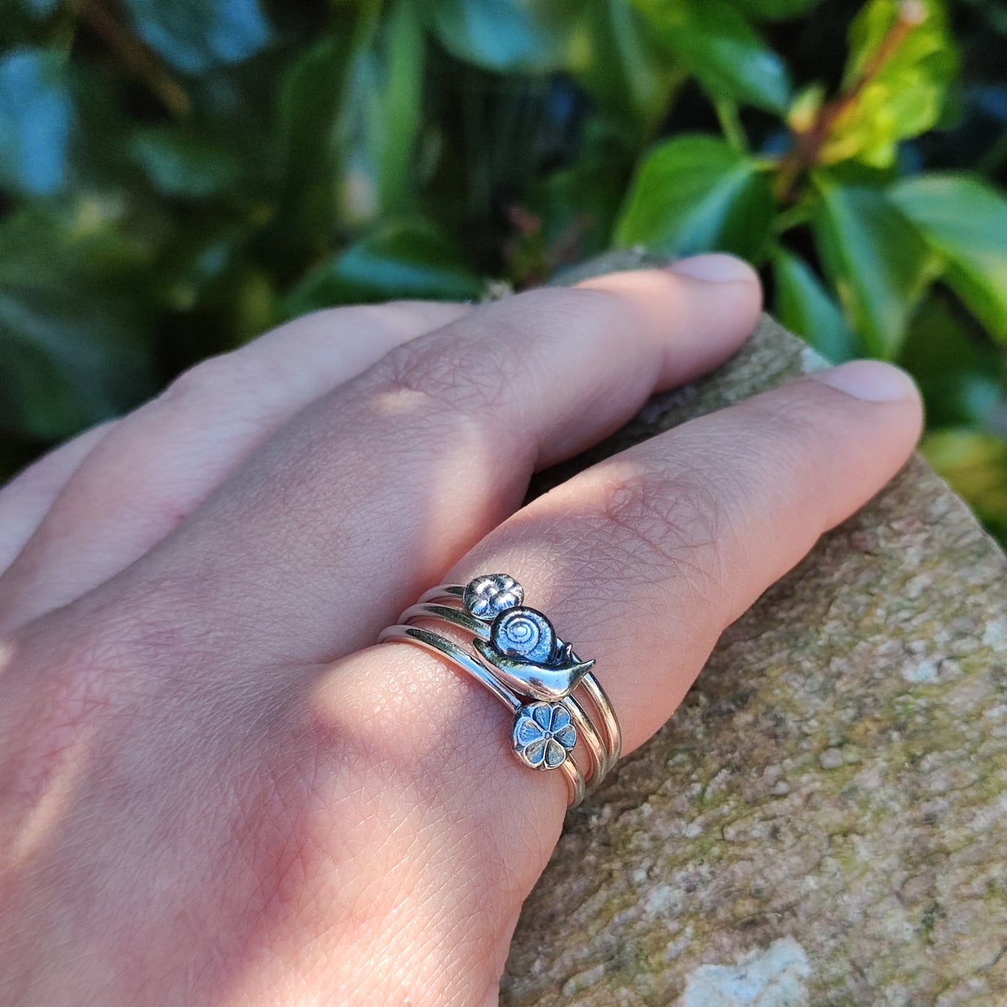 100% recycled silver stacker rings, two with flower motifs and one with a snail, being worn on the index finger. Ivy in the background.