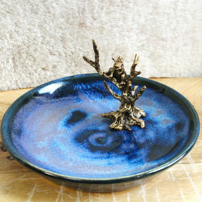 A round blue glazed ceramic dish with a metallic bronze tree branch sculpture standing in the center, set on a light wood surface against a textured neutral background.