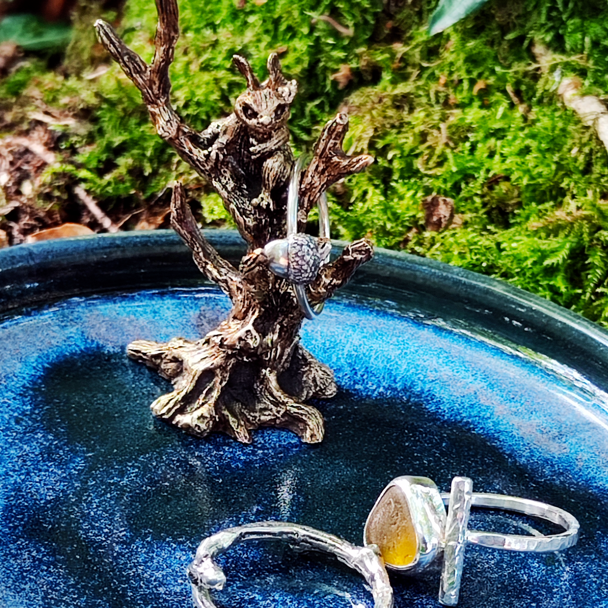 Two silver rings displayed on a small blue ring dish featuring a brass red squirrel sitting in a tree, with a mossy forest background and green leaves in the foreground. One ring has a textured band, and the other features a round yellow stone in a silver setting.